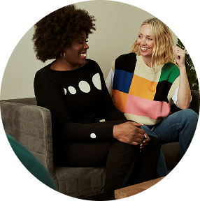 Two women smiling on a couch, chatting in colorful sweaters