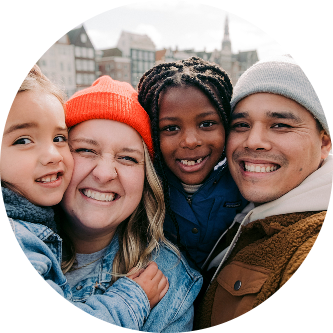 Four friends smiling close together outdoors in winter hats and coats, with city buildings in the background