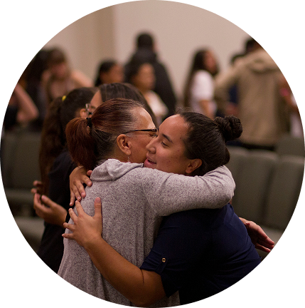 Two people hugging in a crowded indoor gathering, eyes closed, with blurred attendees in the background