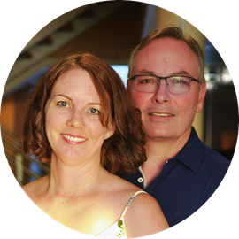 Two smiling adults posing indoors, one in a white floral top and one in a navy shirt with glasses.