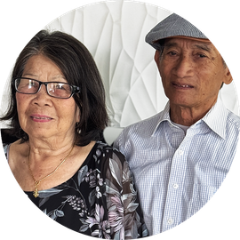 Two people posing together, one wearing glasses and a floral blouse, against a white patterned background