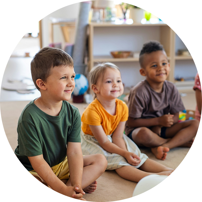 Children sitting cross-legged on the floor in a bright classroom, listening and smiling