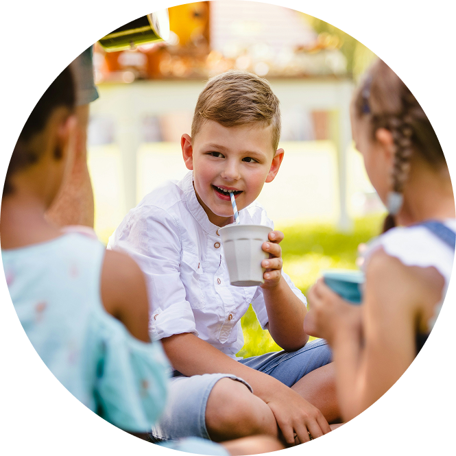 Child drinking from a cup while sitting outdoors with two blurred people in the foreground