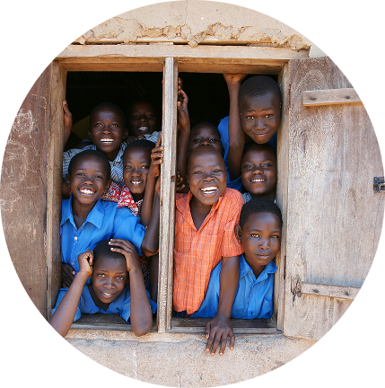 Smiling children in blue and orange shirts looking out of a window in a rustic building