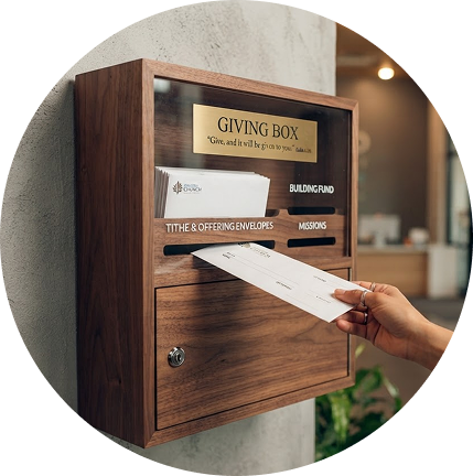 Hand placing an envelope into a wooden giving box mounted on a wall.