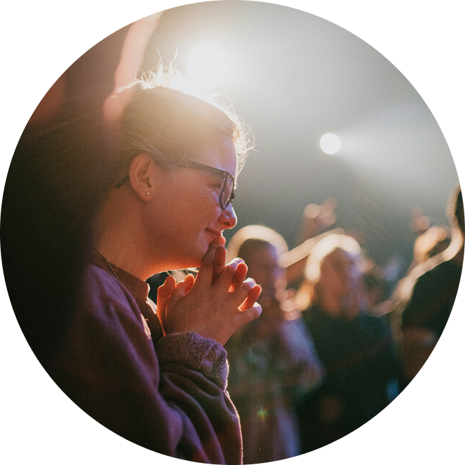 Person in glasses, hands clasped, in bright stage light with a blurred audience behind.
