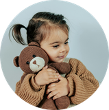 Young child hugging a brown teddy bear, wearing a tan sweater against a light blue background