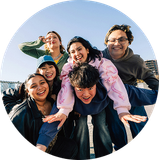 Five friends smiling outdoors in a playful group pose against a blue sky.