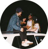 Three people sit and chat on top of a white car at night.