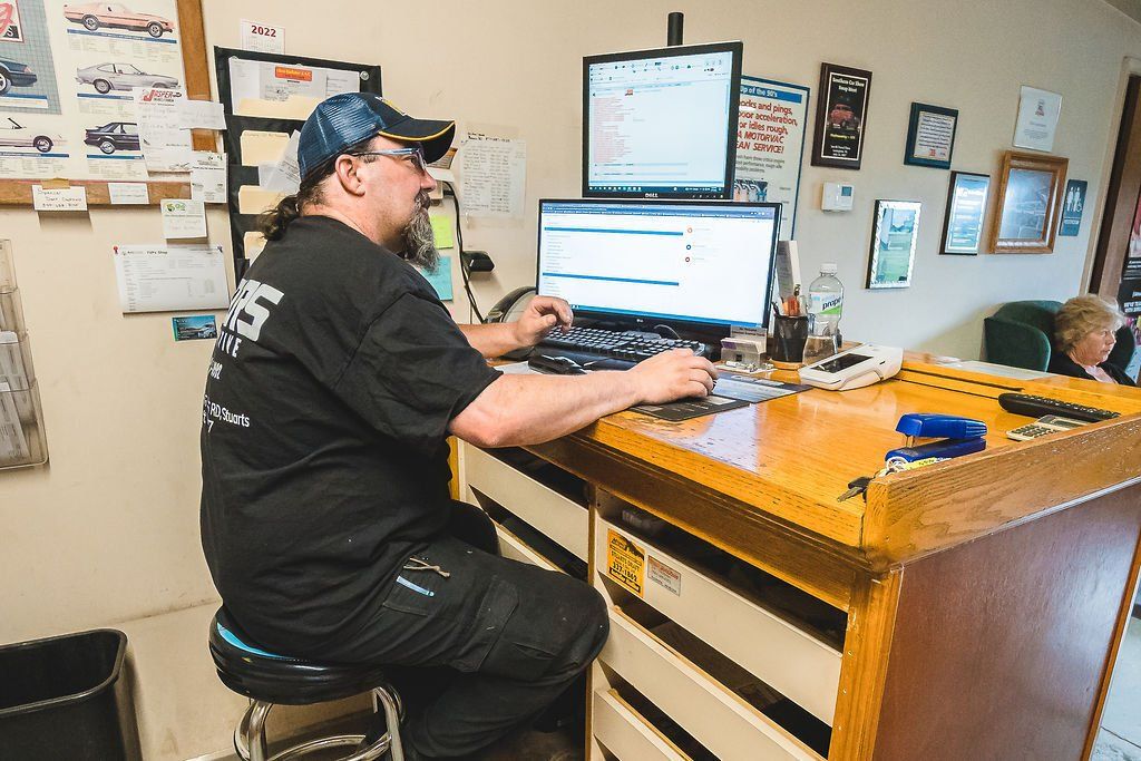 Man in black shirt and cap types at a computer in an office, two monitors visible.