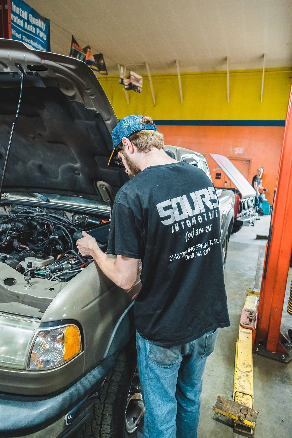 Mechanic working on a car with the hood open in a garage. Wearing a blue cap and a black shirt.