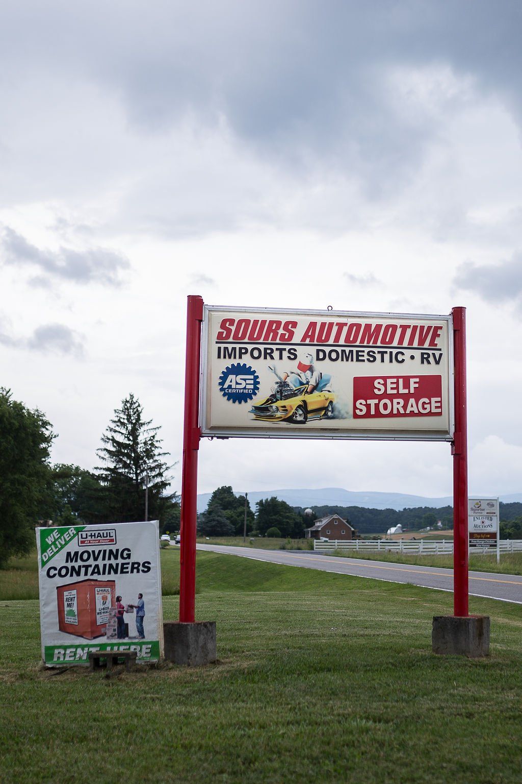 Sign for Sours Automotive, imports and domestic, with self-storage. Red and white sign, cloudy sky.