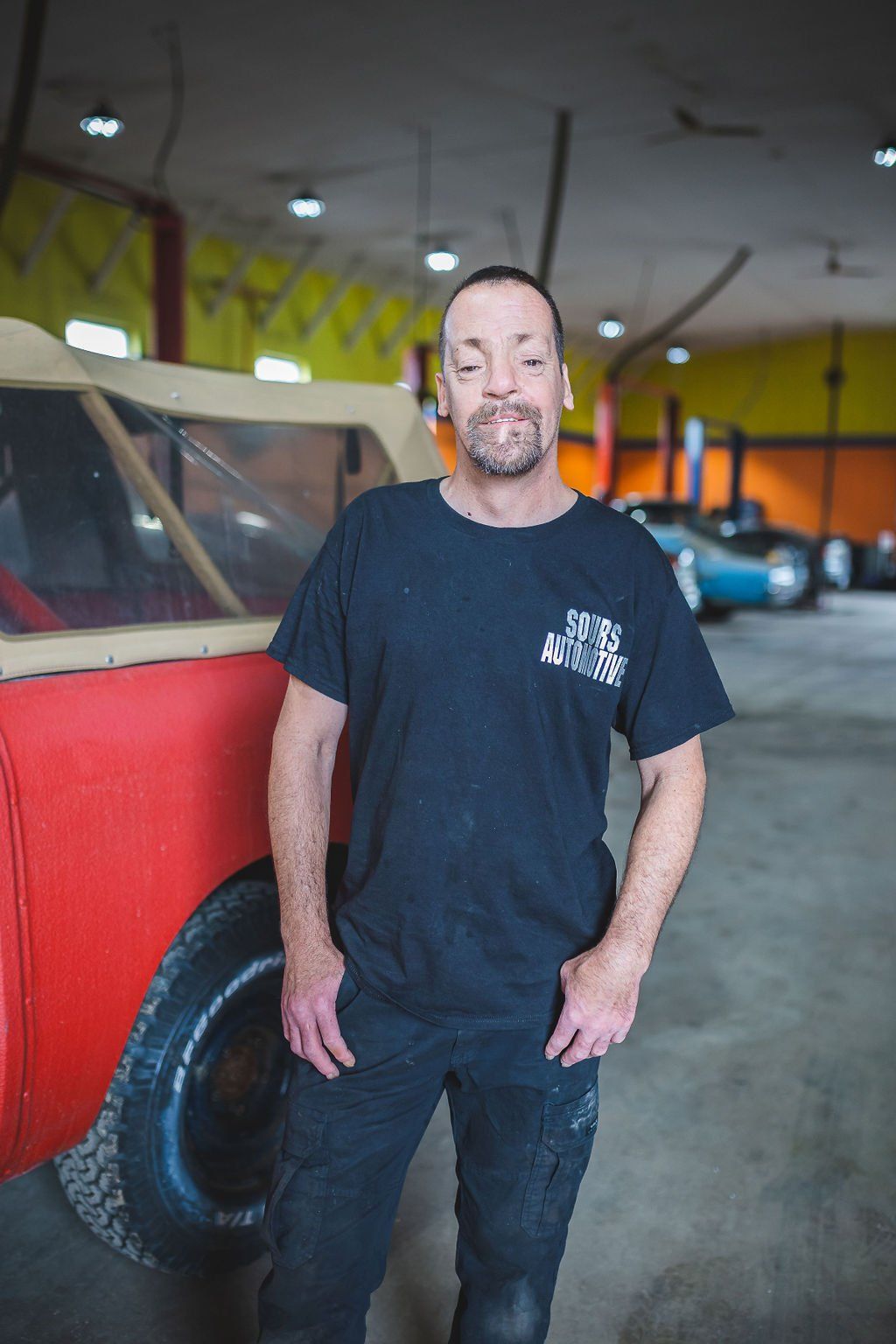 Man in black t-shirt and jeans stands in a garage beside a red car, hand in pocket, looking at camera.