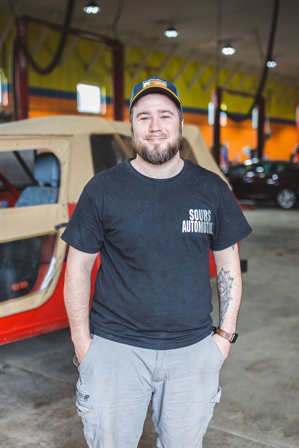 Man in a garage wearing a cap and black shirt with hands in pockets, standing near a red and tan Jeep.
