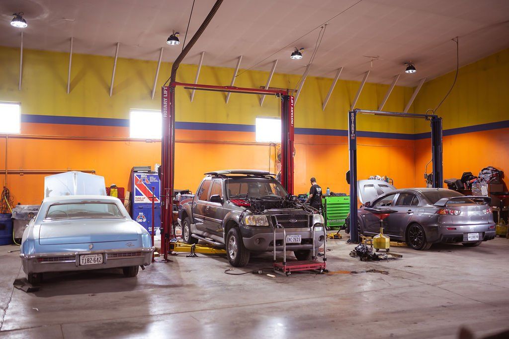 Cars being worked on in a garage. Pickup truck on a lift, classic car, and another car visible.