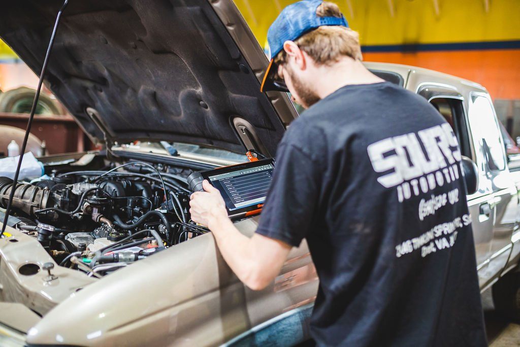 Mechanic in blue hat and black shirt working on a car with the hood open in a garage.