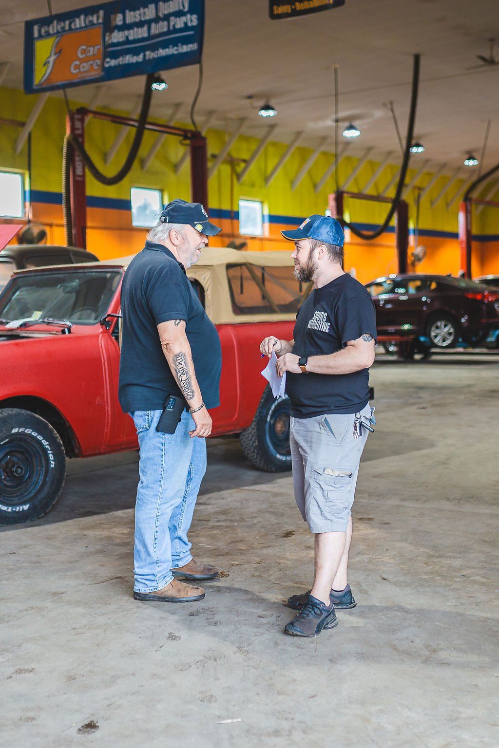 Two men conversing in a garage. Red pickup truck in the background. Overhead service bay.