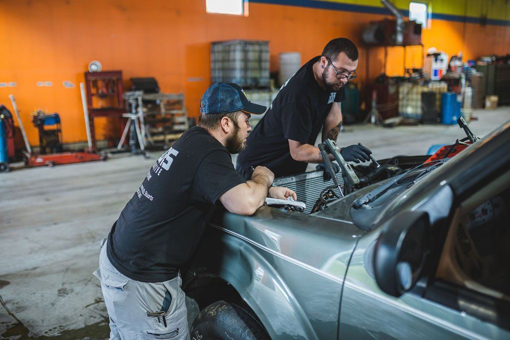Two mechanics working on a car engine in a garage; one leans on the hood.