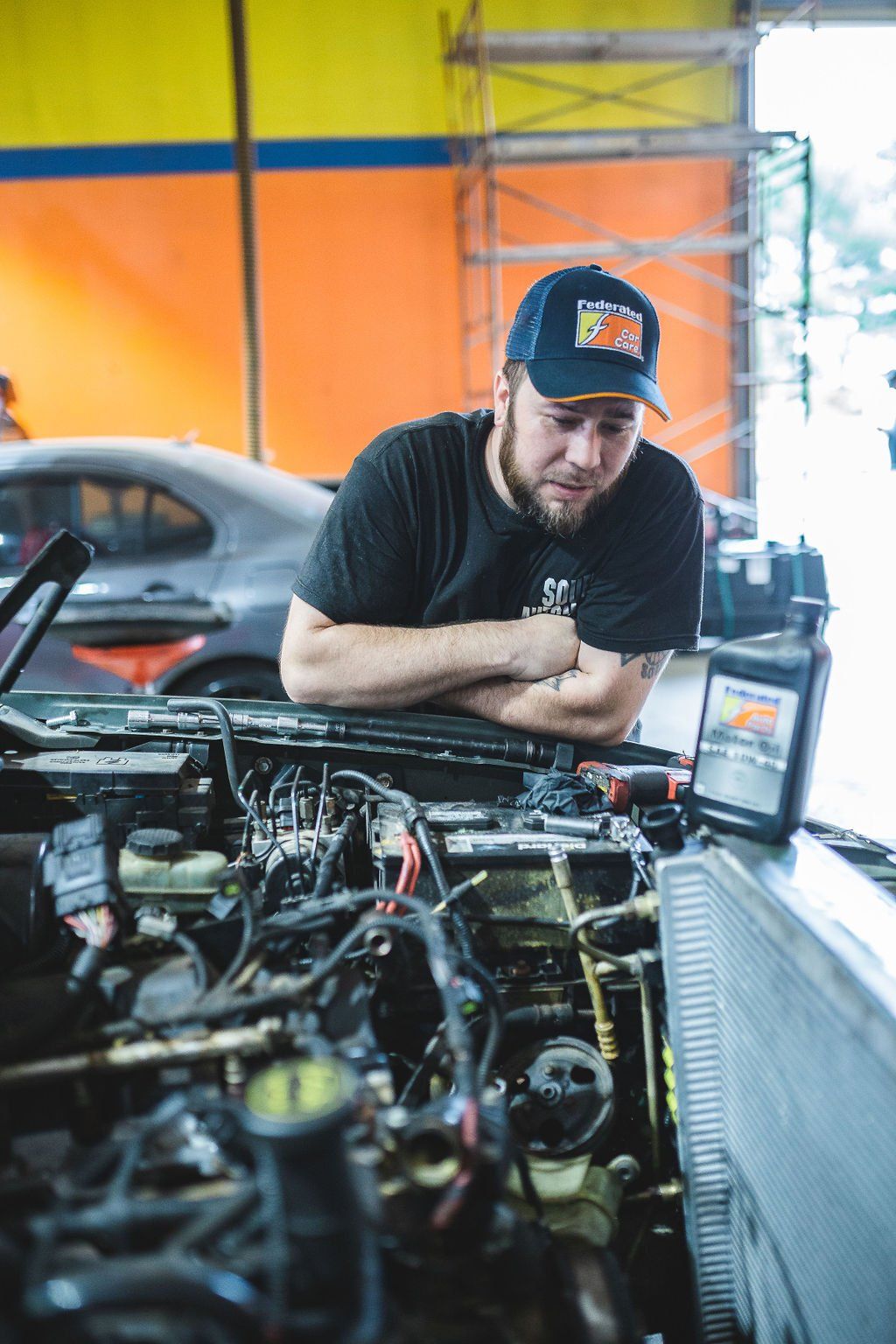 Mechanic examining car engine in shop; wearing cap and looking at manual.