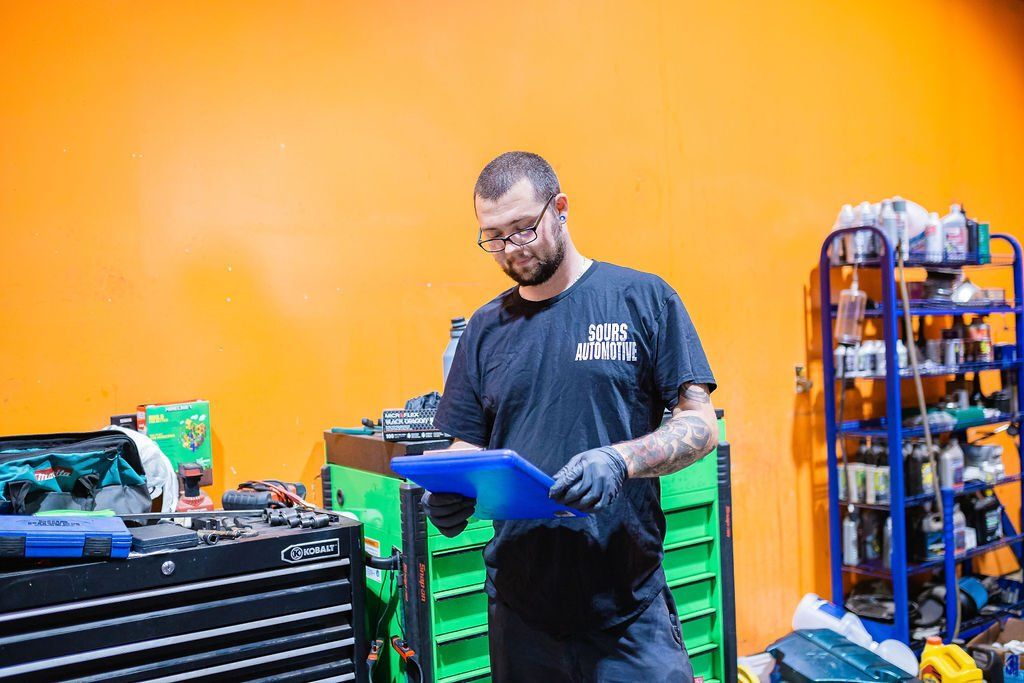 Mechanic in garage, reviewing paperwork. Wearing gloves, looking down, orange wall behind.