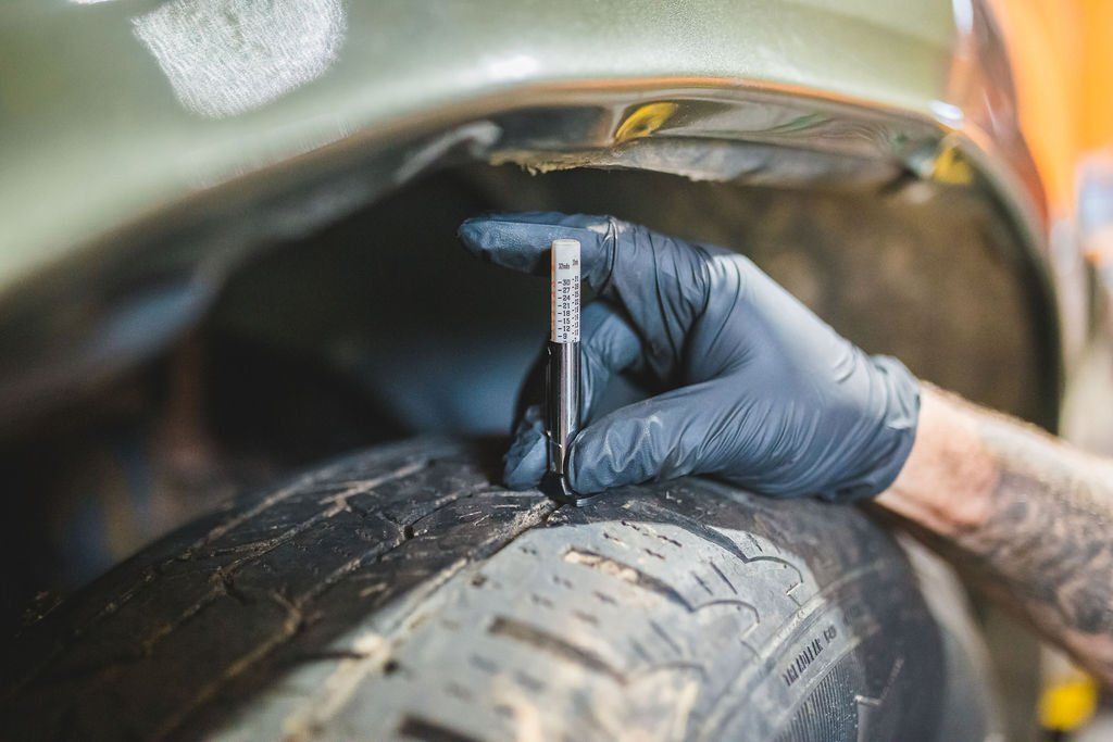 Gloved hand measuring tire tread depth with a gauge in a vehicle's wheel well.
