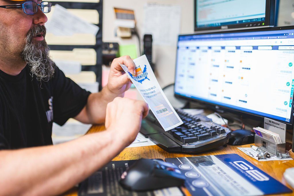Man at a desk looking at a printed document, computer monitor showing schedule.