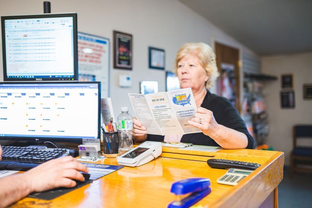 Woman reading brochure at a desk, someone works on a computer nearby. Office setting.