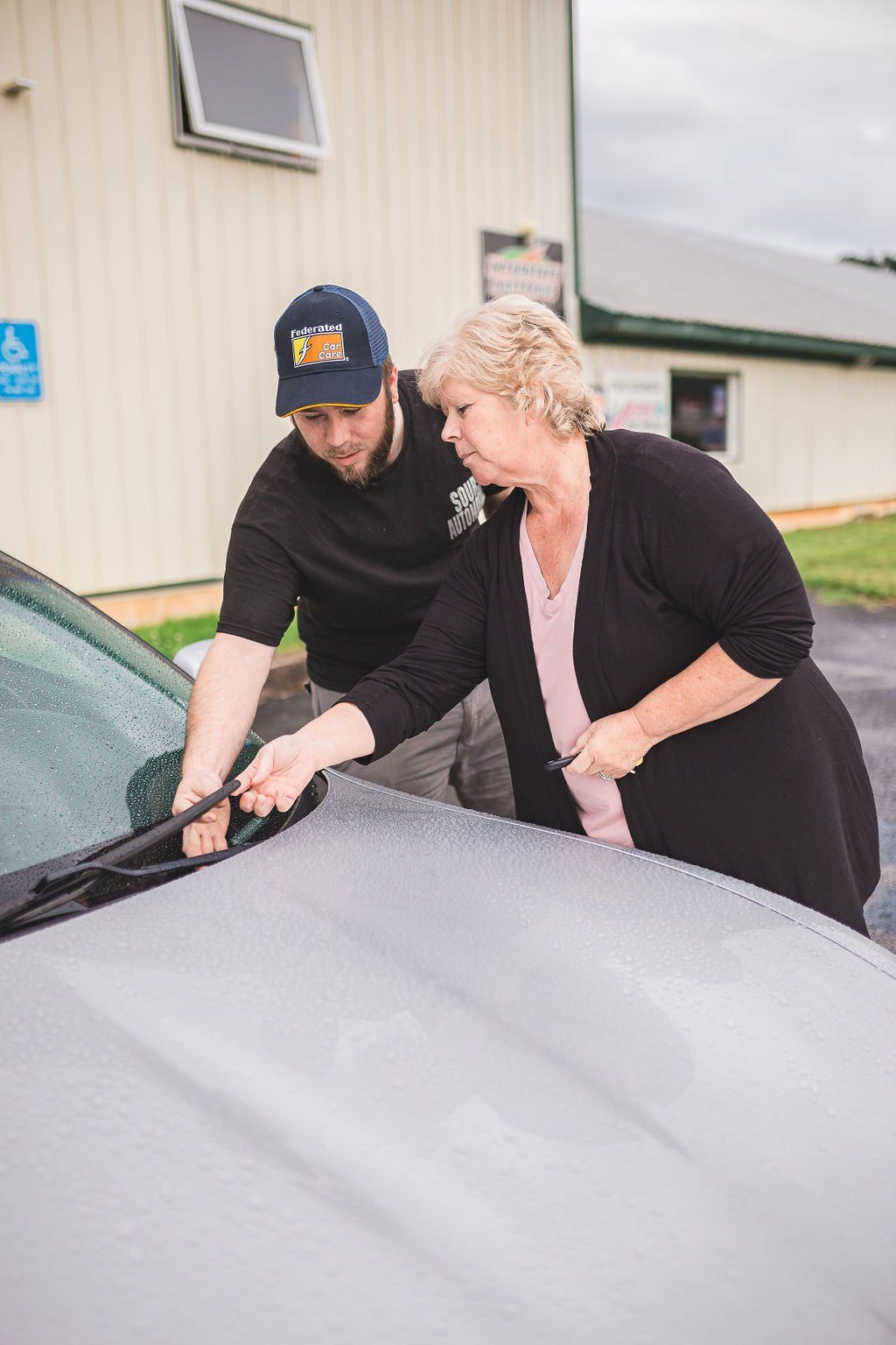 Man and woman adjusting windshield wiper on a silver car outdoors.