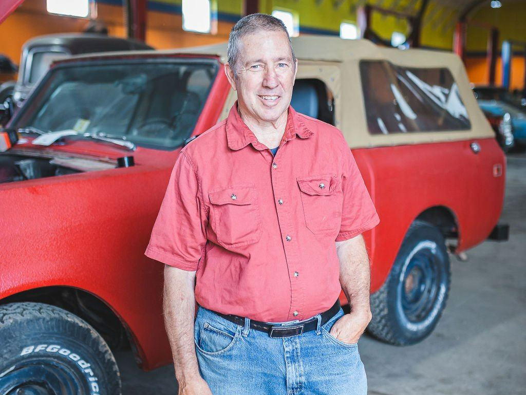 Man in red shirt and jeans stands beside a red vintage SUV, inside a garage.