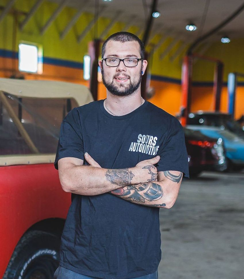 Mechanic with tattoos at auto shop, arms crossed, wearing a black shirt, standing in front of red car and other vehicles.