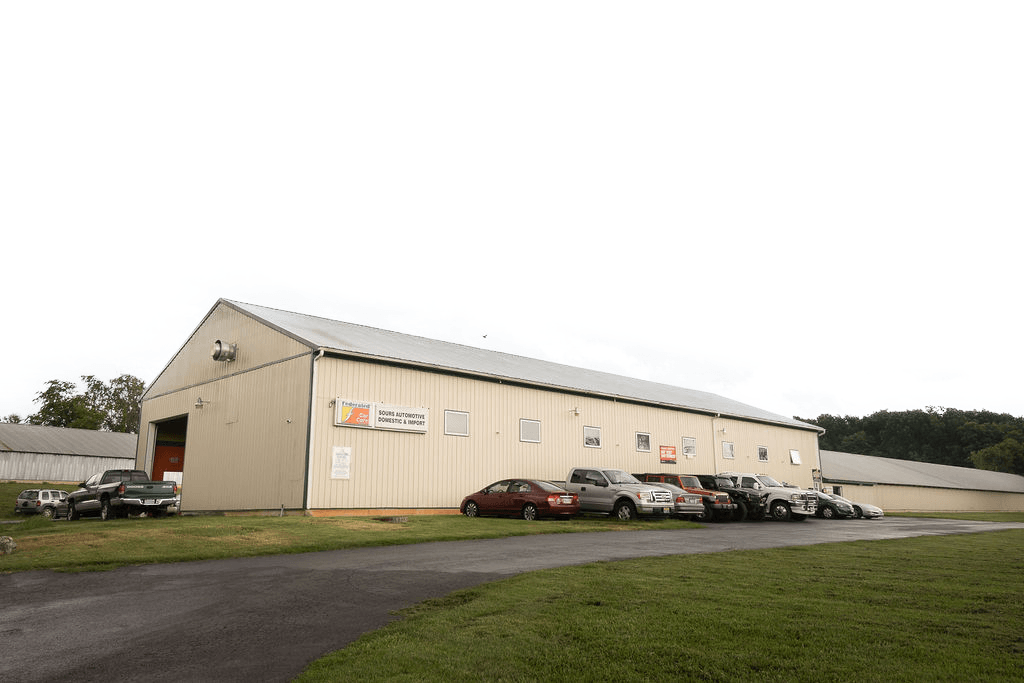 Large, tan metal warehouse with parked cars and a paved driveway, set against a cloudy sky.