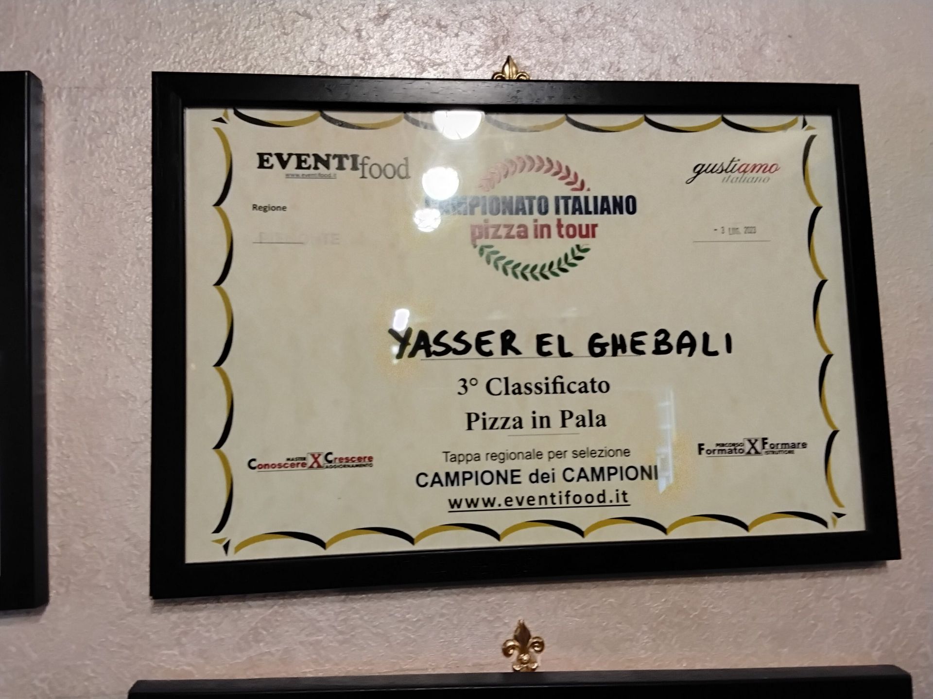 Attestato di riconoscimento incorniciato e appeso al muro, con bordo oro e nero e testo in italiano per Yasser El Emeed.