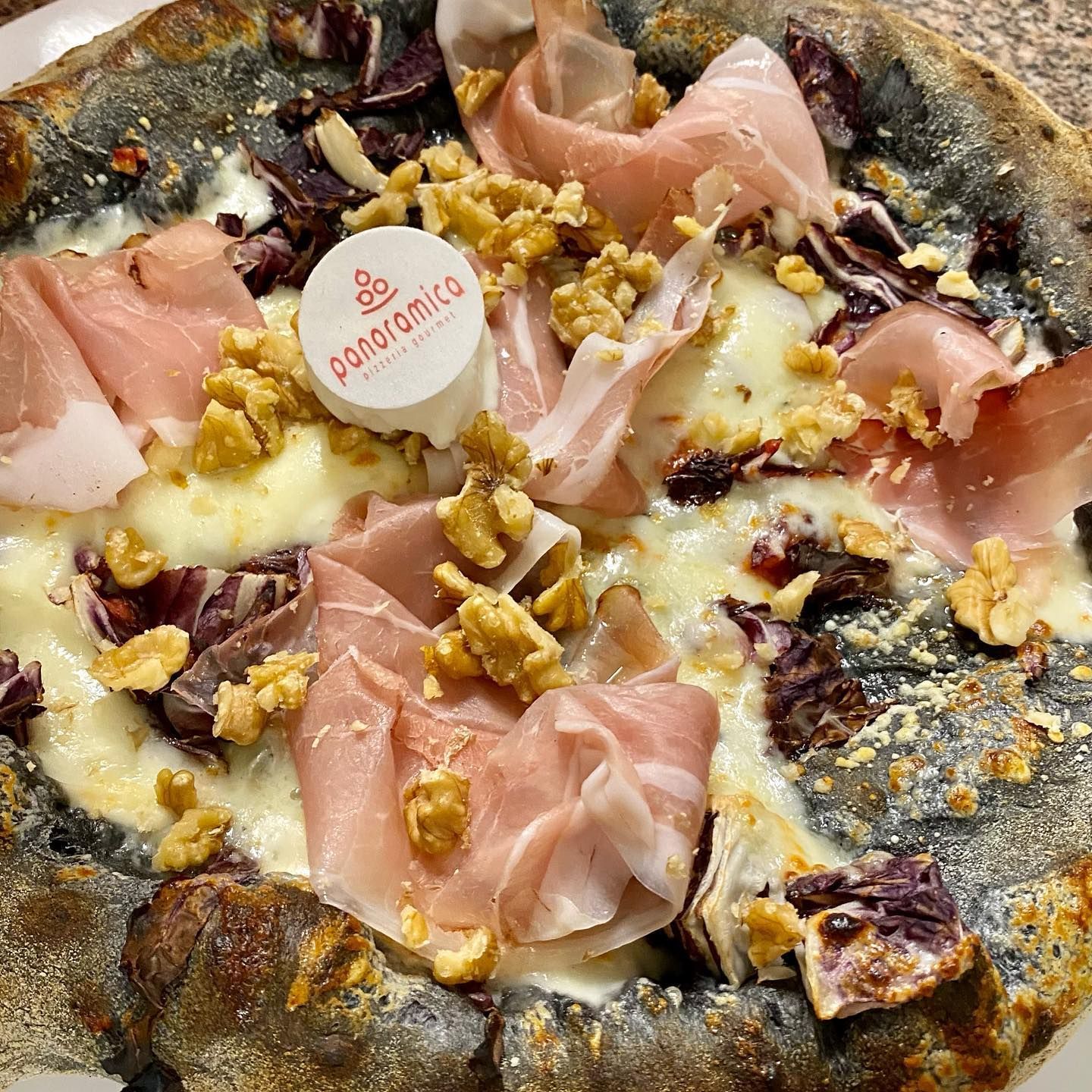 Primo piano di una pizza gourmet condita con prosciutto, formaggio e noci su una base rustica.