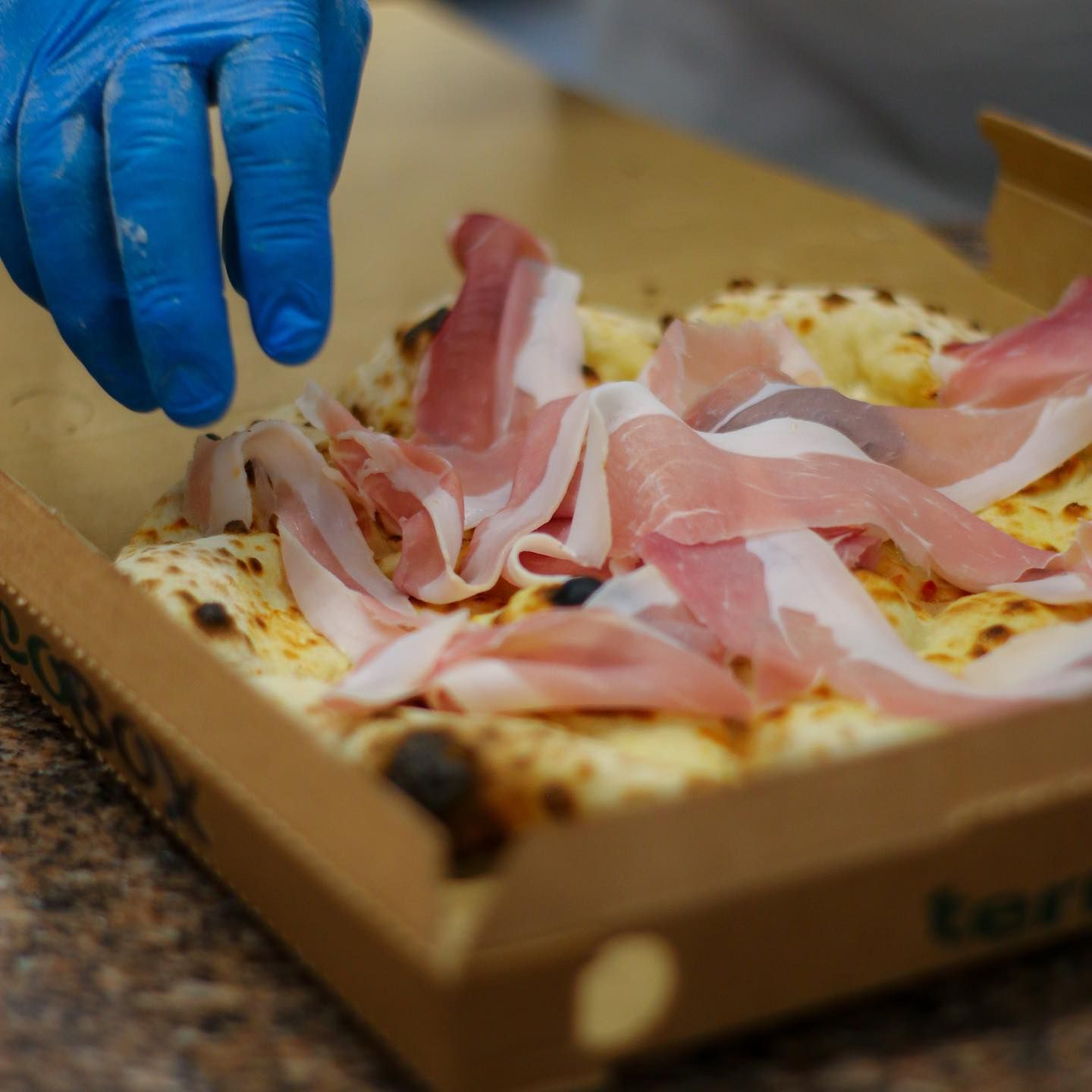 Mano guantata che posiziona fette di prosciutto su una pizza in una scatola di cartone