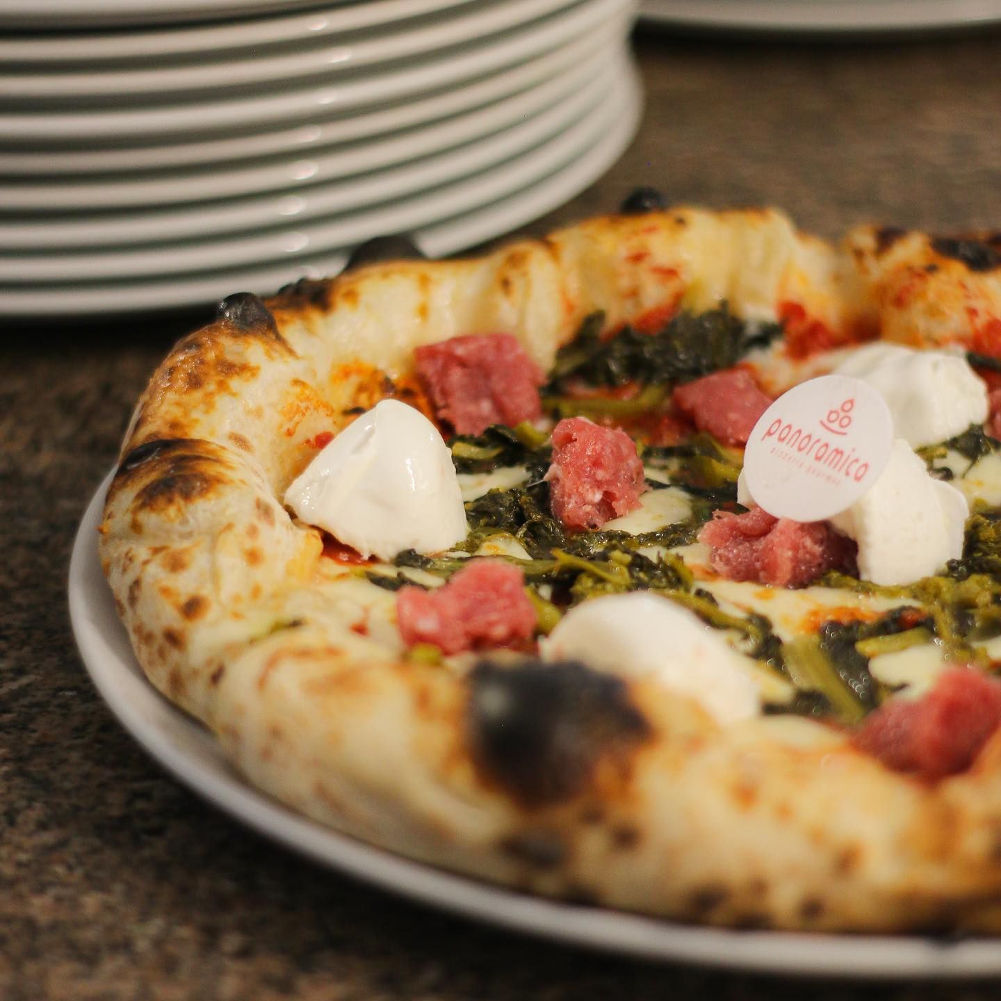 Pizza cotta nel forno a legna con formaggio e carne a cubetti, su un tavolo accanto a piatti impilati.