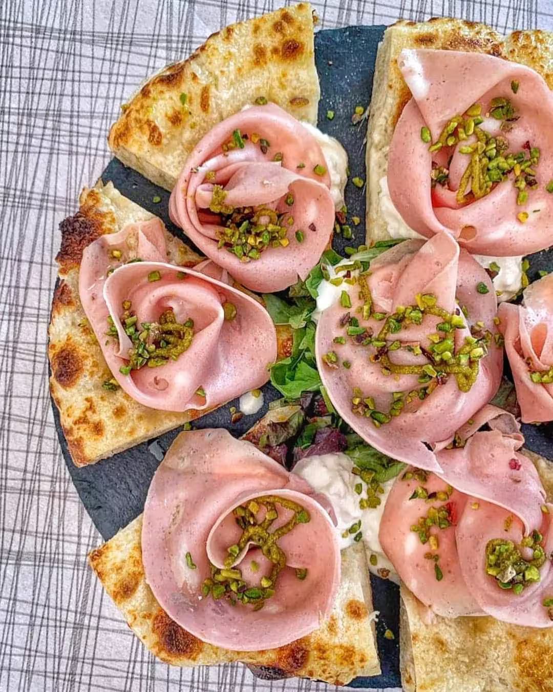 Pizza su focaccia con prosciutto rosa ripiegato e pistacchi tritati, servita su un piatto scuro.