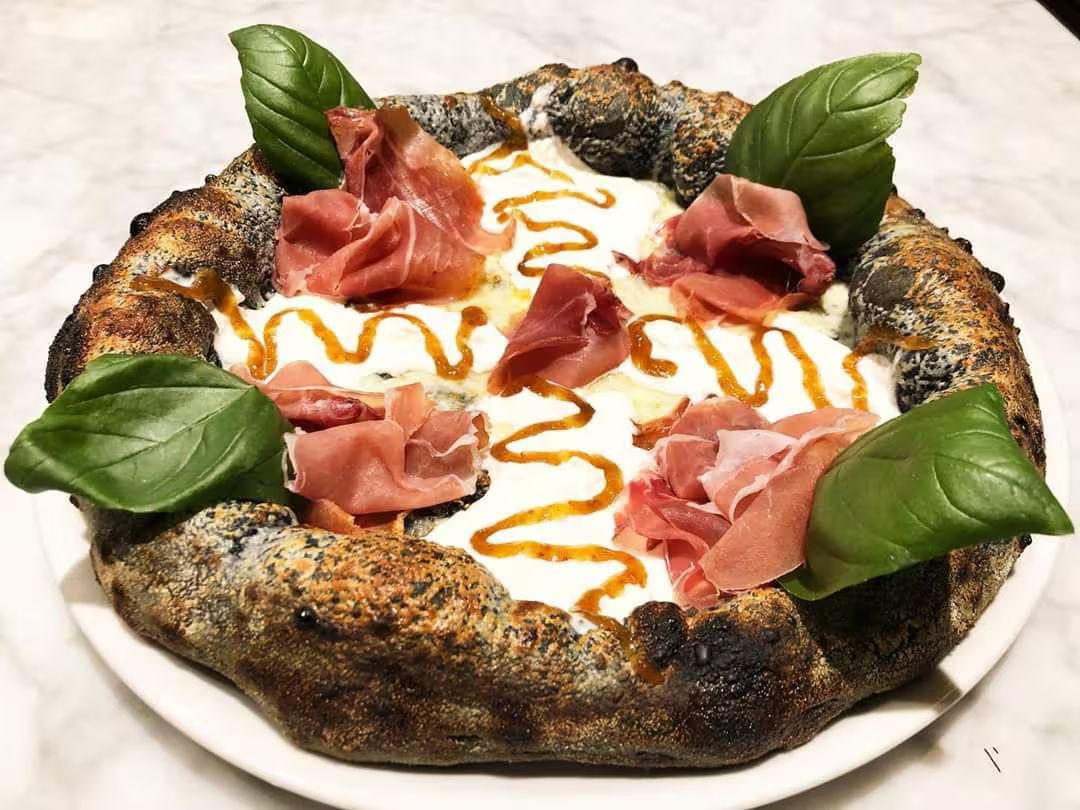 Pizza napoletana condita con foglie di basilico, prosciutto e vortici di salsa su un piatto bianco.