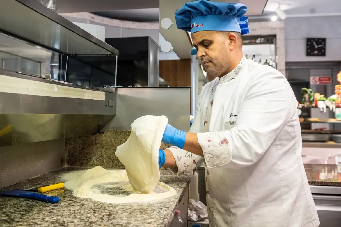 Chef in uniforme bianca e cappello blu versa la farina su un bancone della cucina, con impasto e utensili nelle vicinanze.