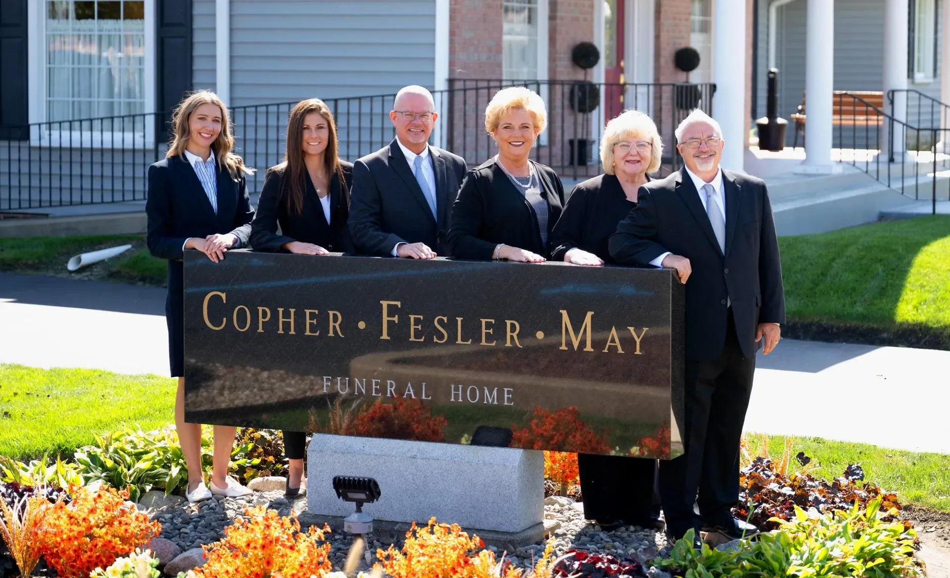 Six professionals stand behind a sign for Gopher Fesler May Funeral Home in front of a building.