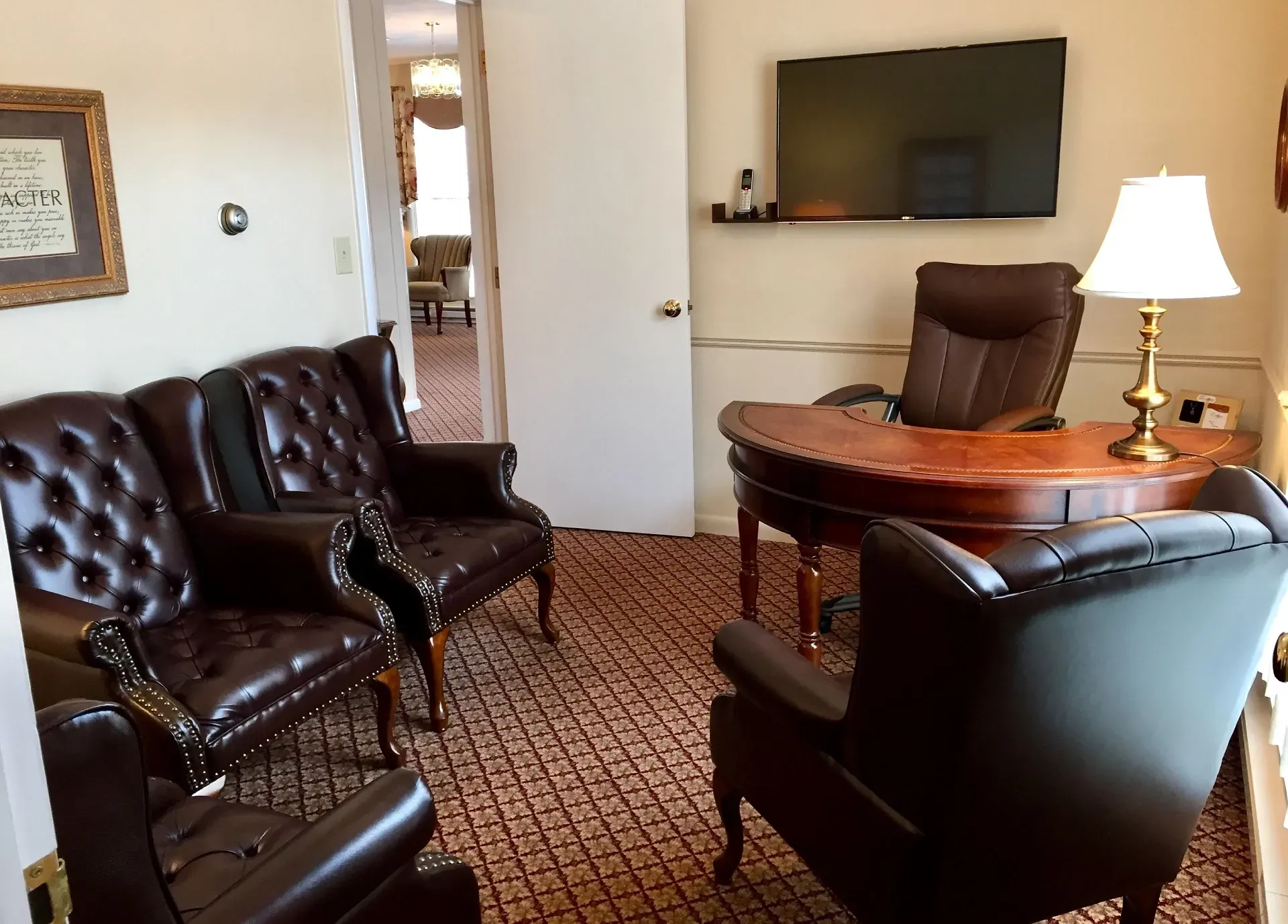 A wood desk with a leather chair, lamp, and television, facing three leather armchairs in an office with patterned carpet.