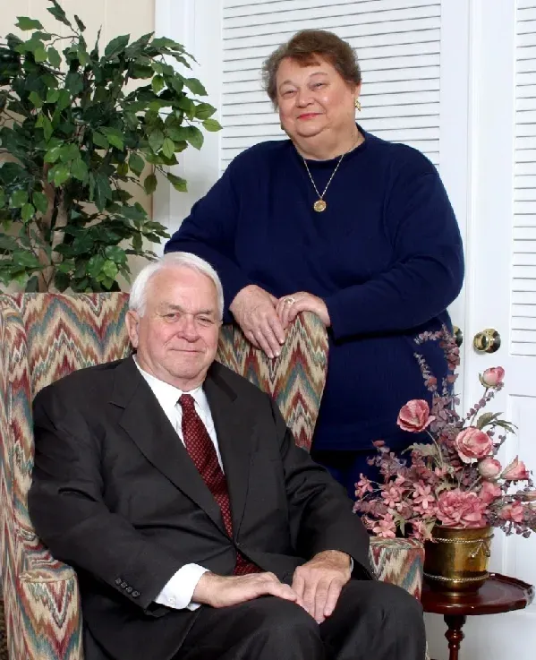 A person in a dark suit sits in a patterned armchair, while another stands behind them in a navy sweater.
