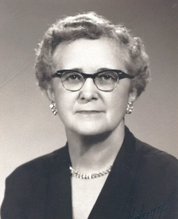 Head-and-shoulders portrait of a person with wavy hair wearing glasses, earrings, a necklace, and a dark jacket.