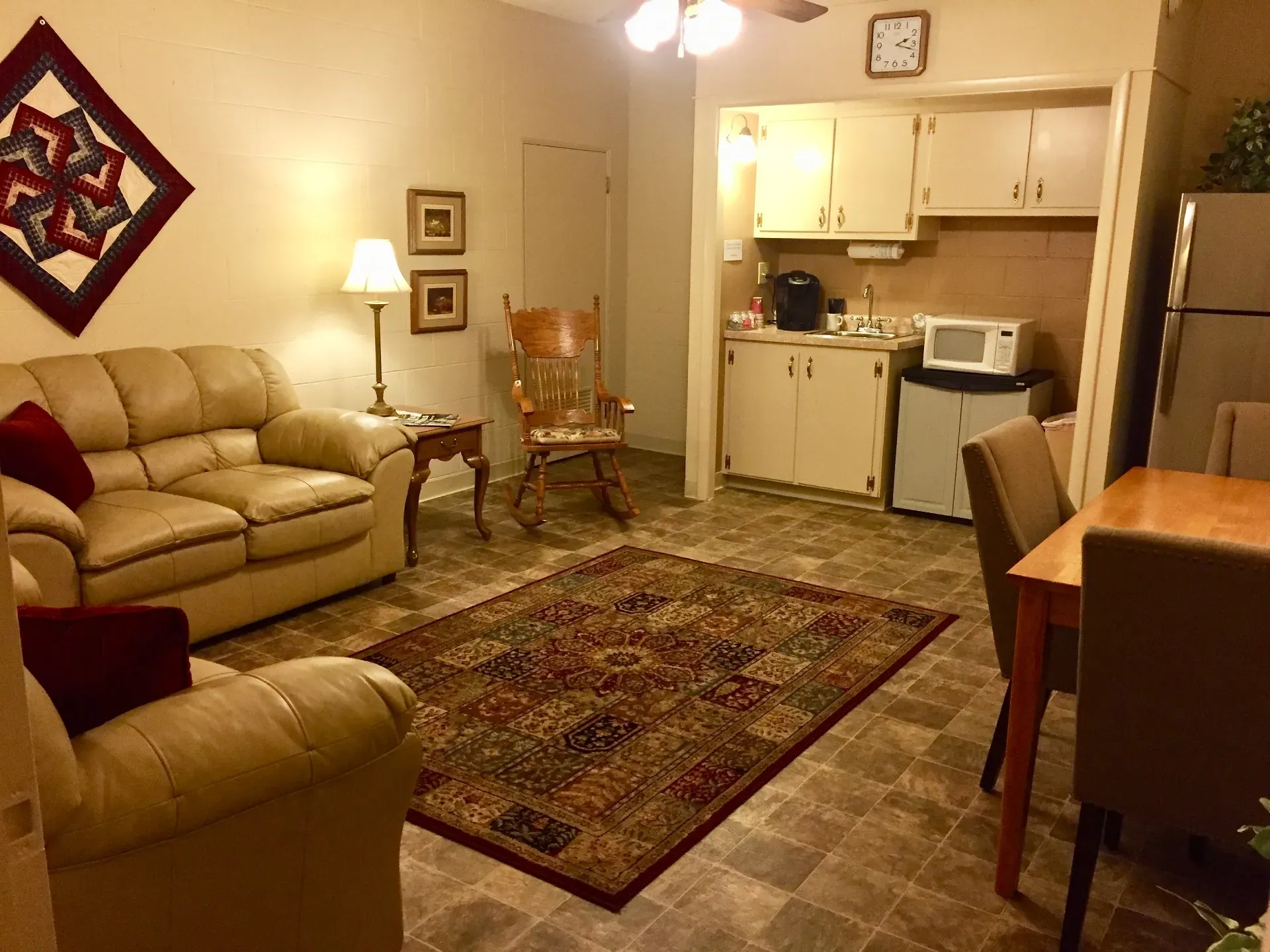 A cozy studio apartment with a leather sofa, patterned rug, wooden rocking chair, and a compact kitchen area.