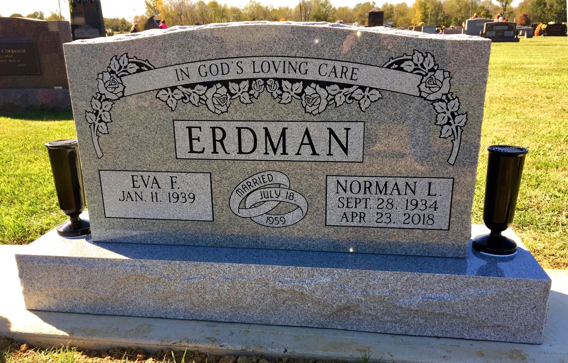 A light grey granite headstone for the Erdman family in a cemetery, featuring names, dates, and floral engravings.