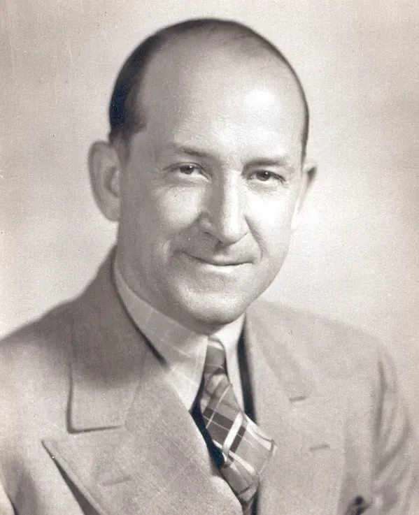 A head-and-shoulders portrait of a smiling person wearing a suit and a patterned tie against a plain background.