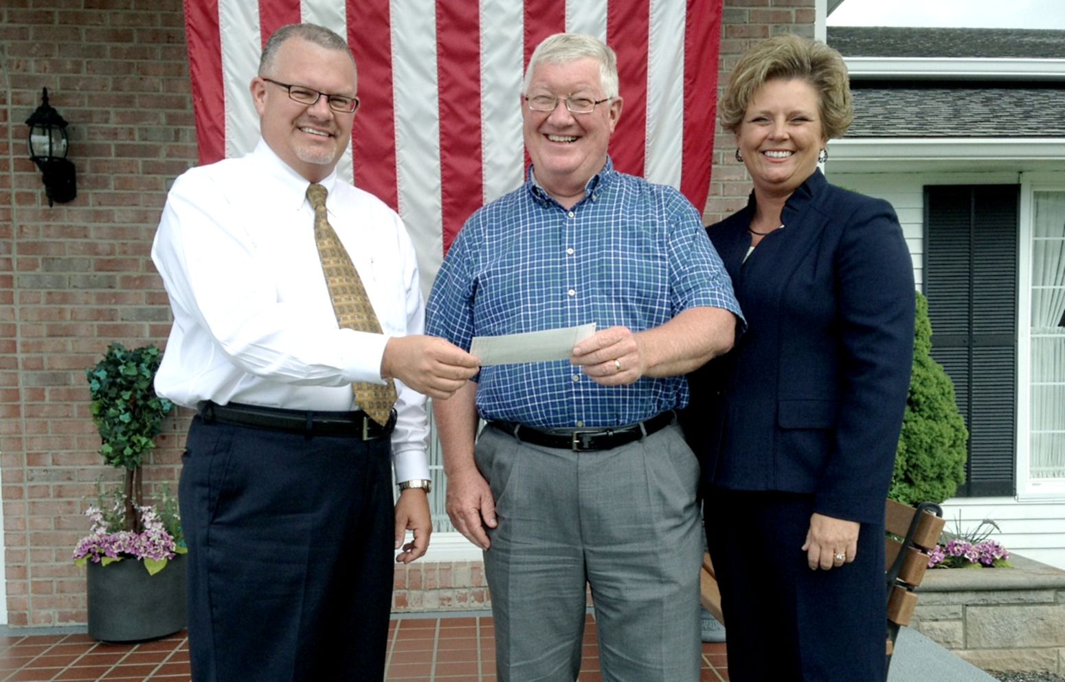 Three people stand in front of an American flag; the center individual holds a paper out to be received by the person left.