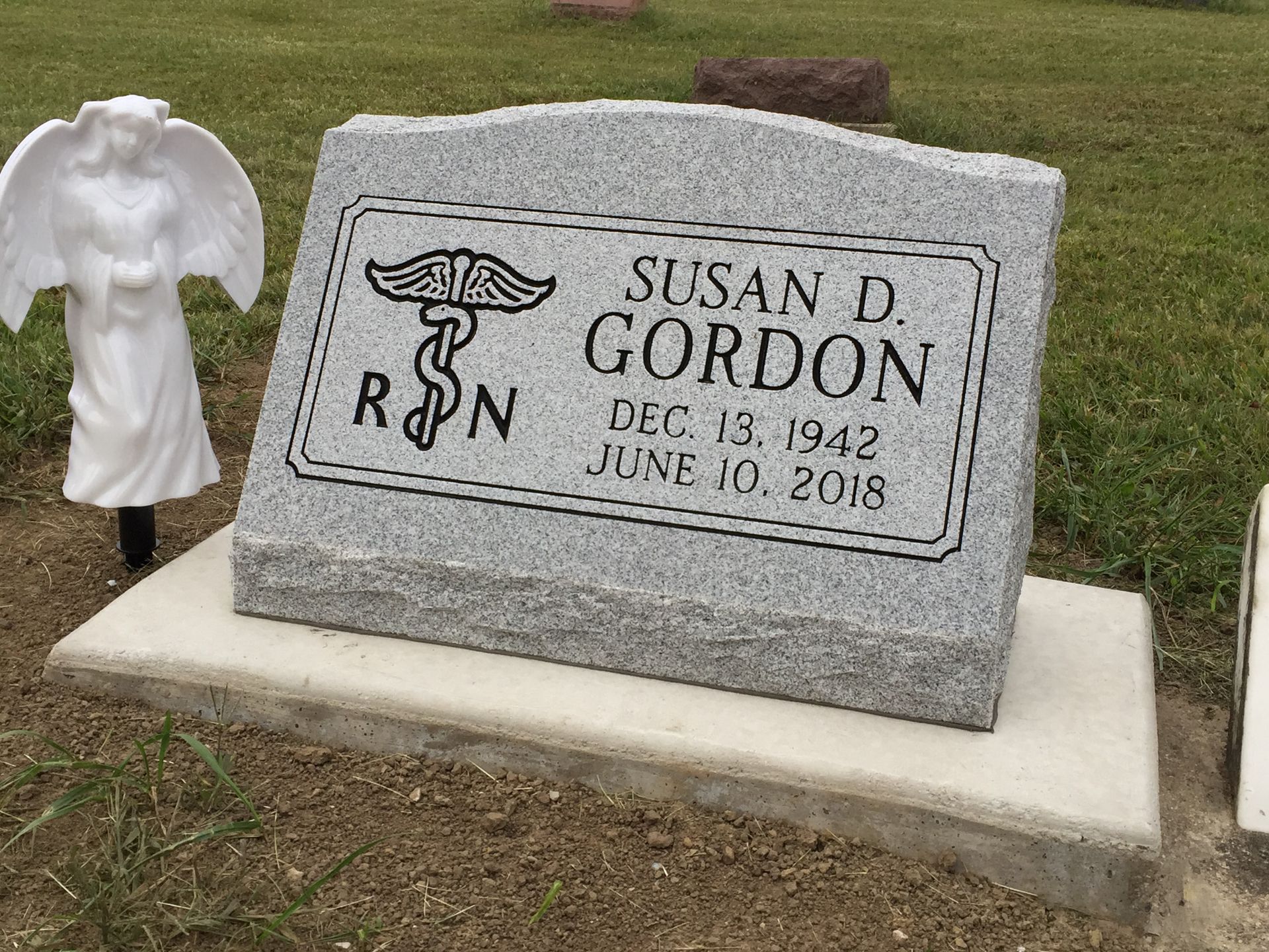 A gray granite headstone for Susan D. Gordon (1942–2018) featuring a nursing emblem, with a small angel statue nearby.