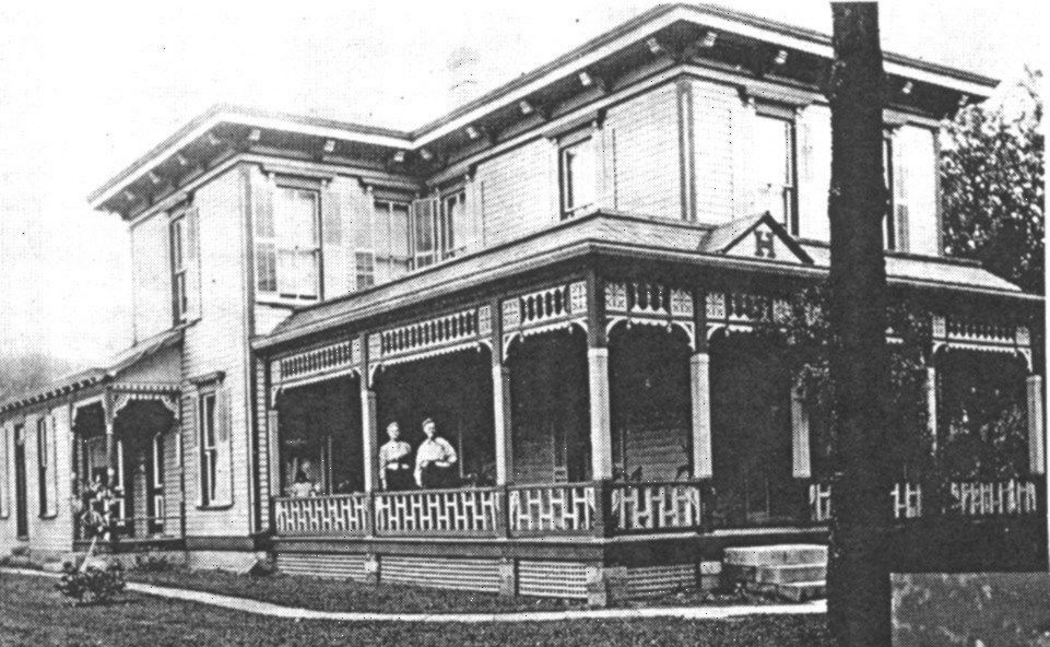 A historic, two-story house with a wrap-around porch and ornate woodwork, featuring two individuals standing on the porch.