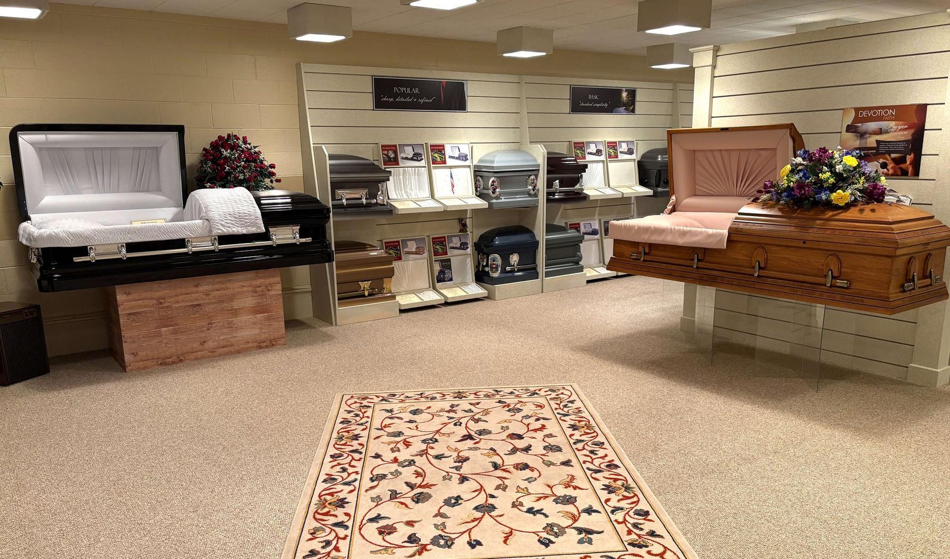A funeral home showroom featuring multiple open and closed caskets on display, with a patterned rug in the center.