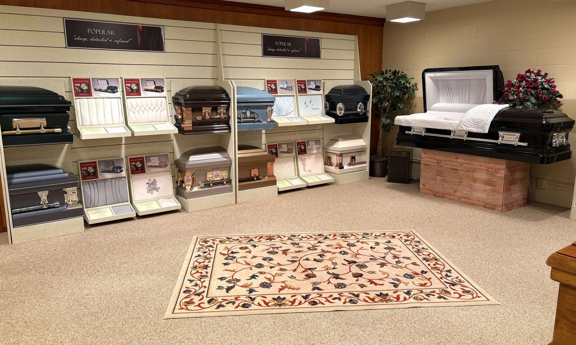 A funeral home showroom featuring multiple casket displays on shelves and a pedestal, with a patterned rug on the floor.